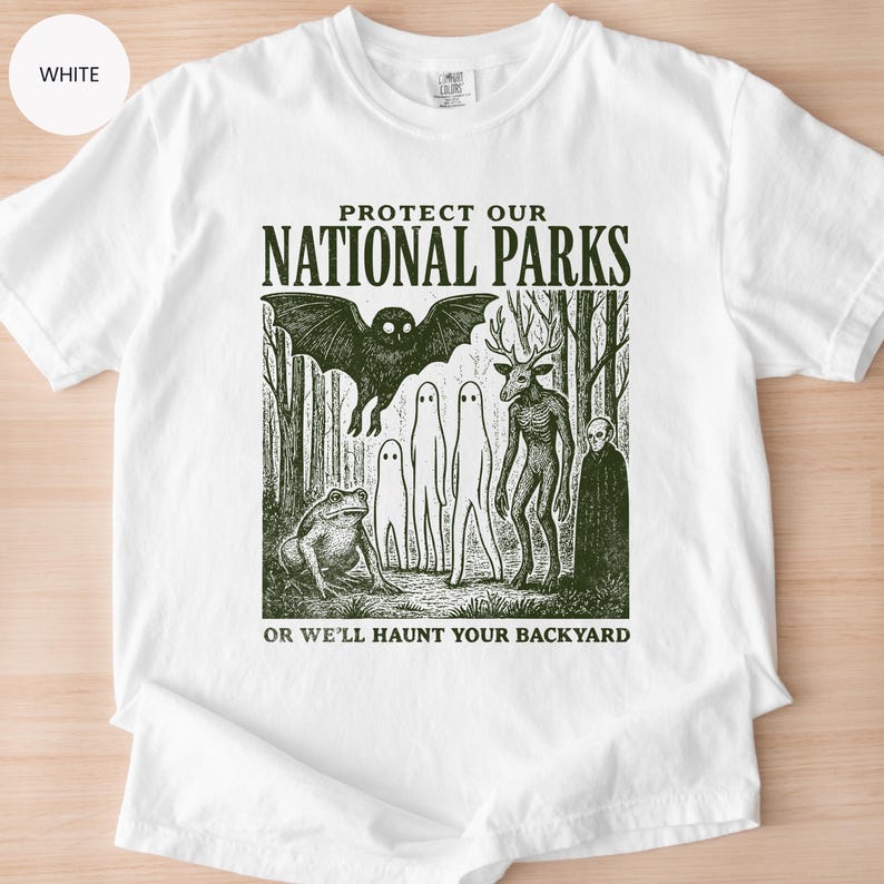 May include: White t-shirt with a forest-themed graphic. The design features a bat, a deer-like creature, and other spooky figures, with the text "PROTECT OUR NATIONAL PARKS OR WE'LL HAUNT YOUR BACKYARD" in a bold font. The shirt is made of a soft material.