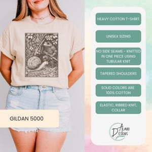 K&ouml;nnte beinhalten: Ein beiges T-Shirt mit einer Vintage-Illustration eines Frosches, der auf einer Schnecke reitet. Das Shirt ist ein Gildan 5000 und besteht aus schwerem Baumwollstoff. Es hat eine Unisex-Gr&ouml;&szlig;en, keine Seitenn&auml;hte, ein Schlauchgewebe, konische Schultern und einen elastischen Rippstrickkragen.