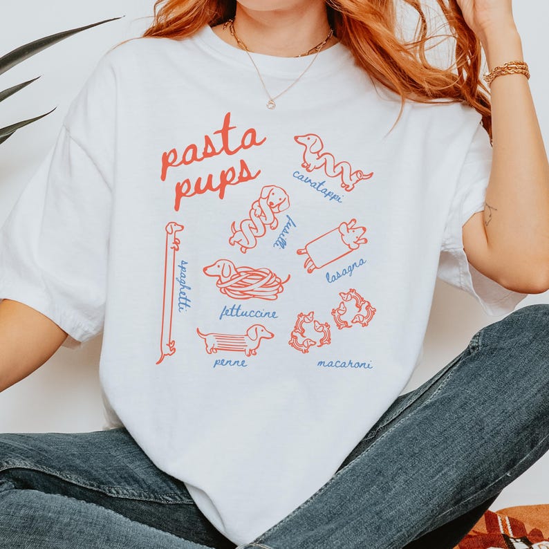 May include: White t-shirt with the text "pasta pups" in red and blue. The shirt features illustrations of dachshund dogs representing different pasta shapes, including spaghetti, fettuccine, and lasagna.