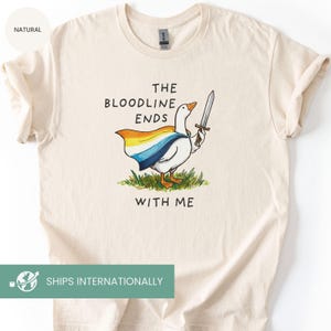 May include: Cream-colored t-shirt featuring a cartoon goose with a sword, wearing a cape with rainbow colors. The text "THE BLOODLINE ENDS WITH ME" is printed above and below the goose illustration. The shirt is made of a soft material.