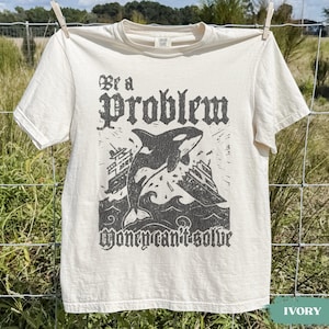 May include: An ivory short-sleeve t-shirt featuring a dark gray graphic print of an orca jumping between two boats. The text above the orca reads "Be a Problem" and the text below the orca reads "Money can't solve" in a stylized gothic font.