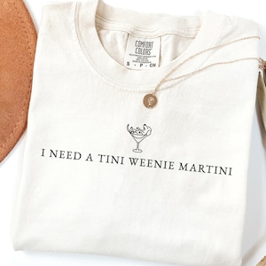 May include: Cream-coloured t-shirt with the text "I NEED A TINI WEENIE MARTINI" printed in black. The shirt has a martini glass graphic and a gold necklace. A brown hat and dried palm leaves are in the background.