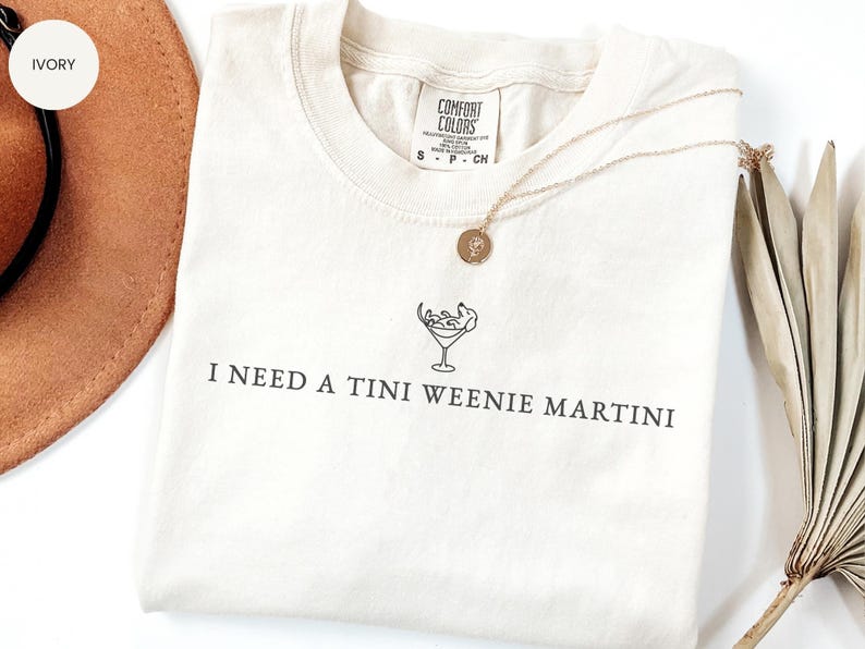 May include: Cream-coloured t-shirt with the text "I NEED A TINI WEENIE MARTINI" printed in black. The shirt has a martini glass graphic and a gold necklace. A brown hat and dried palm leaves are in the background.