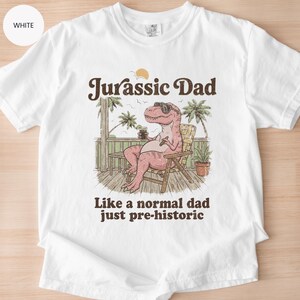 May include: White t-shirt with the text "Jurassic Dad" above an illustration of a relaxed pink Tyrannosaurus Rex wearing sunglasses and holding a drink. The text "Like a normal dad just pre-historic" is below the illustration.
