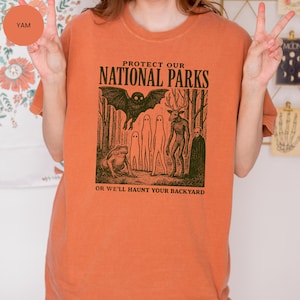 May include: Orange t-shirt featuring the text "PROTECT OUR NATIONAL PARKS" above a forest creature illustration. The text "OR WE'LL HAUNT YOUR BACKYARD" is below the graphic. The shirt is a warm, autumnal colour.
