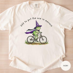 May include: Ivory-colored t-shirt featuring a green frog riding a bicycle, wearing a wizard hat and cape with the asexual pride flag colors. The text "Off to put the ace in menace" is printed above the frog.