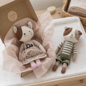 May include: Two handmade stuffed animal dolls: a cat in a floral dress with the name "Lia" and a dog in striped shirt and shorts. The cat is in a cardboard box with a sticker that says "Especially for you". The dolls are on a white surface.