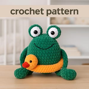 May include: A green crochet frog toy with large white-rimmed eyes and a smiling face, embracing an orange duckling. The words "crochet pattern" are above the frog. The toy is on a wooden surface.