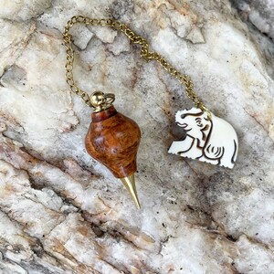 May include: A pendulum with a wooden body and brass tip, connected by a gold chain to a white elephant charm. The wooden body has a unique grain pattern. The elephant charm is carved with details.