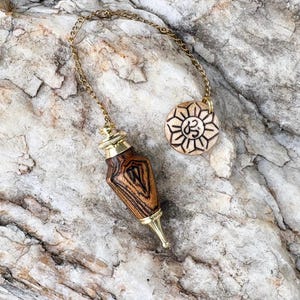 May include: A wooden pendulum with a gold chain and accents, featuring a teardrop-shaped body with a detailed wood grain pattern. A round, engraved charm with a sunburst design and the Om symbol is also attached. The items are set against a textured, light-colored stone surface.