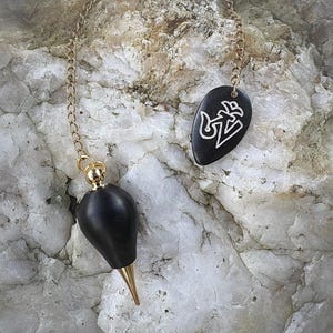 Puede incluir: Un péndulo negro con cadena y detalles dorados. El péndulo tiene un peso en forma de lágrima con punta dorada. Un dije negro de forma ovalada con un diseño blanco cuelga de la cadena. El fondo es una piedra clara y texturizada.