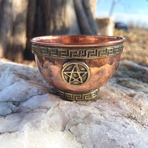 Puede incluir: Un pequeño cuenco de cobre con un diseño de pentagrama y un borde de clave griega. El cuenco está sentado sobre una roca blanca.