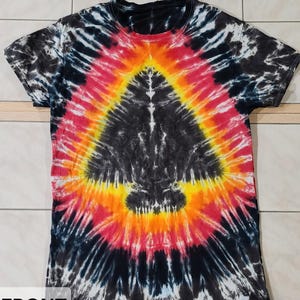 Pu&ograve; includere: Una maglietta tie-dye con un motivo a spirale nero, rosso, arancione e giallo. La maglietta ha un girocollo e maniche corte. La parola "FRONT" &egrave; visibile in basso a sinistra.