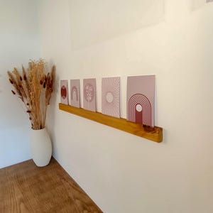 Peut inclure: Une étagère en bois présente cinq impressions d'art avec des motifs abstraits dans des tons de rose et de bordeaux. Les impressions présentent des formes géométriques et des motifs floraux. Un vase blanc avec des fleurs séchées est posé sur une surface en bois.