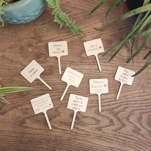 Op de afbeelding: Een set van tien houten plantenstokjes met verschillende berichten erop geschreven. De berichten zijn: "Maman qui déchire", "Merci Maitresse", "Viens on sème", "Arrose-moi si tu peux", "Mamie d'amour", "Si je crève ce sera ta faute", "Parfois je me trouve moche alors je pense à toi et ça va mieux", "Pas la peine de marroser morue".