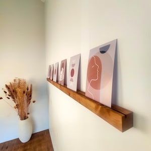 Peut inclure: Une étagère en bois présente une série d'estampes d'art avec des motifs abstraits et une esthétique minimaliste. Les estampes présentent des couleurs sourdes comme le pêche, le beige et le bordeaux, avec des formes géométriques et des illustrations de profils féminins. Un vase blanc avec des fleurs séchées est dans le coin.