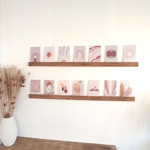 Puede incluir: Colección de láminas de arte abstracto en tonos rosa, melocotón y marrón, expuestas en dos estantes de madera. Las láminas presentan varios diseños geométricos e inspirados en la naturaleza, incluyendo soles, arcos y formas orgánicas. Un jarrón blanco con flores secas está en primer plano.