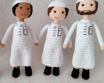 May include: Four crocheted dolls wearing white robes and white hats. The dolls have brown and black hair and brown eyes. The dolls are standing in a row on a white surface.