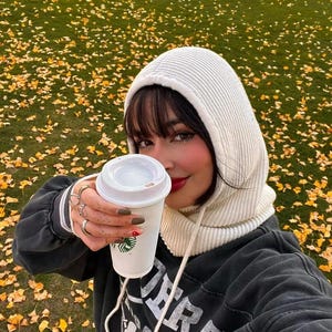 Könnte beinhalten: Eine Person hält einen Starbucks-Kaffeebecher. Der Becher ist weiß mit dem Starbucks-Logo. Die Person trägt einen weißen Kapuzenschal und einen schwarzen Sweatshirt. Der Hintergrund ist eine grüne Wiese mit gelben Blättern.