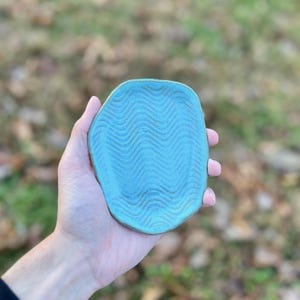 May include: A handmade, turquoise ceramic dish with a wavy pattern. The dish is irregularly shaped and appears to be held in a person's hand. The background is blurred, with hints of green and brown.
