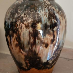 May include: A large, round ceramic vase with a glossy finish. The vase features a blend of dark brown, beige, and light blue colors, creating a unique, textured appearance. The base of the vase is a lighter, earthy tone.
