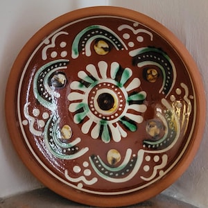 May include: A round, terracotta-colored ceramic plate with a detailed floral design. The plate features a central flower motif with white petals, a brown center, and green accents. The rim is a darker brown.