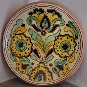 May include: A round ceramic plate with a floral design. The plate features a cream-colored background with yellow, green, and black floral patterns. The rim is brown with a white and brown border. The plate is likely handmade and suitable for decorative use.