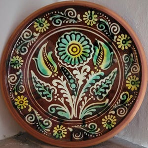 May include: A decorative ceramic plate with a brown background and a floral design. The central flower is turquoise with a yellow center, surrounded by white, green, and yellow floral accents. The rim is brown.