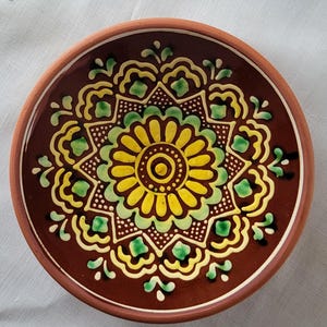 May include: A round, decorative ceramic plate with a brown rim and a dark brown center. The center features a floral design in yellow, green, and white, with a detailed, symmetrical pattern. The plate is likely handmade.
