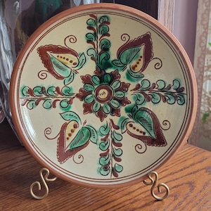 May include: A decorative ceramic plate with a cream-colored background and a brown rim. The plate features a symmetrical floral design in shades of brown, green, and yellow. The plate is displayed on a gold-colored metal stand.