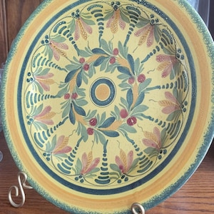 May include: A round ceramic plate featuring a yellow center with a dark blue circle. The surface is decorated with a repeating pattern of green leaves, pink flower-like shapes, and small dark blue dots, finished with a wide green and yellow rim.