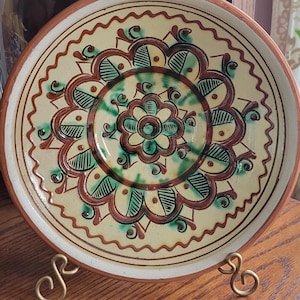 May include: A decorative ceramic plate with a floral design in shades of brown, green, and cream. The plate has a scalloped edge and is displayed on a gold-colored metal stand. The plate's design features a central flower motif.