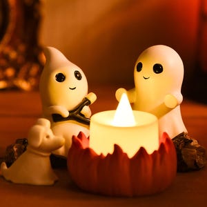 May include: A Halloween-themed decorative scene featuring two smiling ghost figurines, a small dog figurine, and a candle with a flame. The candle sits in a red pumpkin-shaped holder, creating a warm, inviting ambiance.
