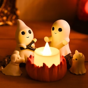 Op de afbeelding: Een Halloween-display met kleine, witte spookfiguren met zwarte ogen en lachende gezichten. Een brandende kaars staat in een rode pompoenvormige houder. Er is ook een klein wit hondenbeeldje aanwezig.