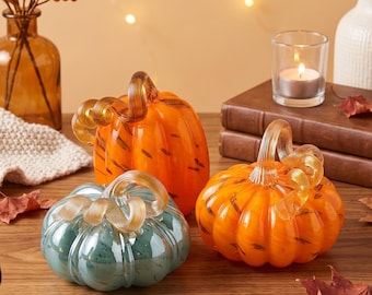 May include: Three glass pumpkins in orange and teal colors, each featuring a clear, spiraled glass stem. The pumpkins have a ribbed, glossy surface and are arranged together on a wooden surface, showcasing their distinct rounded shapes.