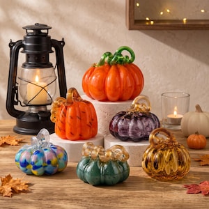 Op de afbeelding: Een verzameling van zes decoratieve glazen pompoenen in diverse kleuren, waaronder oranje, paars, amber, groen en een meerkleurig blauw en geel patroon, elk met een gedraaide glazen steel, geplaatst naast een zwarte lantaarn en een kaars.