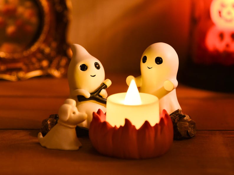 May include: A Halloween-themed decorative scene featuring two smiling ghost figurines, a small dog figurine, and a candle with a flame. The candle sits in a red pumpkin-shaped holder, creating a warm, inviting ambiance.