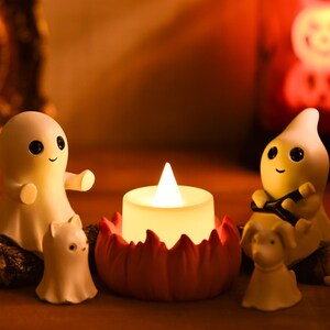 May include: A Halloween-themed display featuring a glowing candle surrounded by smiling ghost figurines and small animal figures. The candle sits in a red, petal-shaped holder. The figurines are white with black eyes and smiling faces.