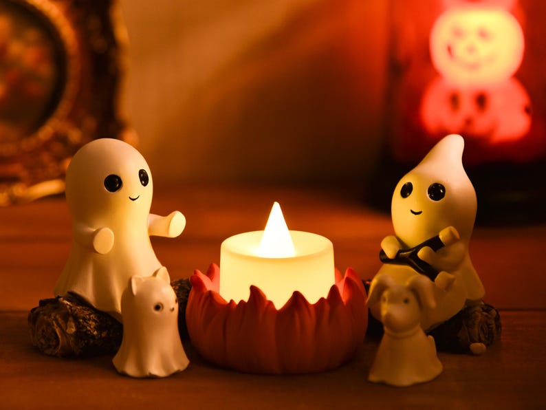 May include: A Halloween-themed display featuring a glowing candle surrounded by smiling ghost figurines and small animal figures. The candle sits in a red, petal-shaped holder. The figurines are white with black eyes and smiling faces.
