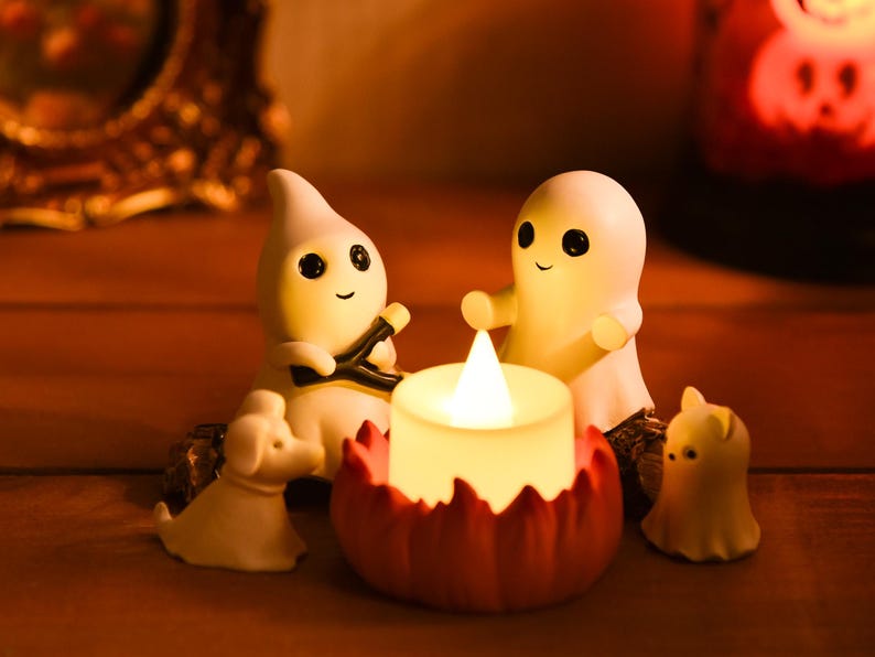 May include: A Halloween-themed display featuring small, white ghost figurines with black eyes and smiling faces. A lit candle sits in a red pumpkin-shaped holder. A small white dog figurine is also present.