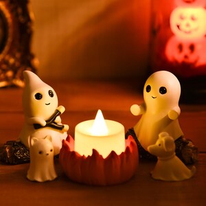 May include: A Halloween-themed display featuring small, white ghost figurines with black eyes and smiling faces. A lit candle sits in a red pumpkin-shaped holder, and a glowing pumpkin lantern is in the background. The scene is set on a wooden surface.