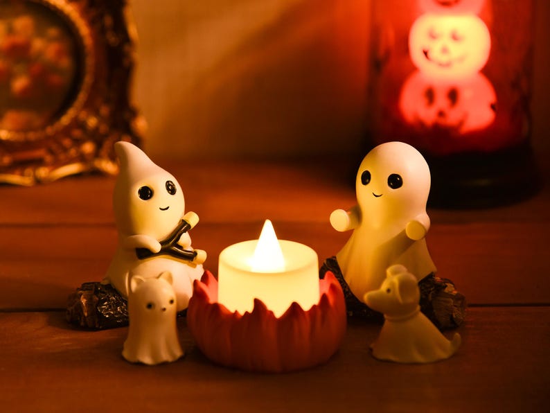 May include: A Halloween-themed display featuring small, white ghost figurines with black eyes and smiling faces. A lit candle sits in a red pumpkin-shaped holder, and a glowing pumpkin lantern is in the background. The scene is set on a wooden surface.