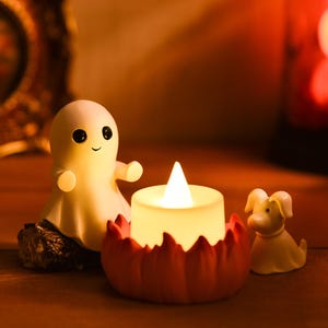 May include: A Halloween-themed display featuring a smiling ghost figurine, a pumpkin-shaped candle holder with a lit candle, and a small dog figurine. The scene is set on a wooden surface with warm lighting, creating a cozy atmosphere.