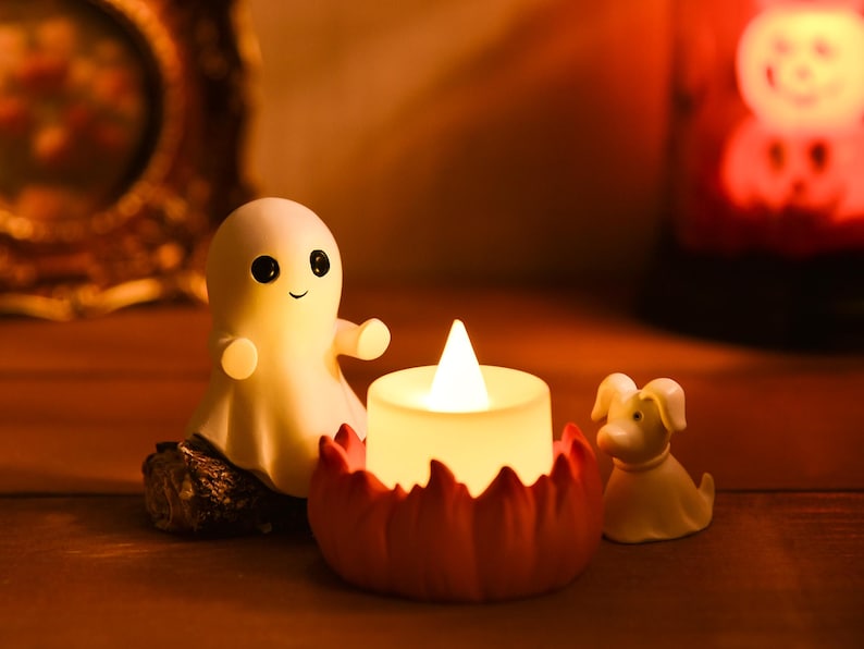May include: A Halloween-themed display featuring a smiling ghost figurine, a pumpkin-shaped candle holder with a lit candle, and a small dog figurine. The scene is set on a wooden surface with warm lighting, creating a cozy atmosphere.