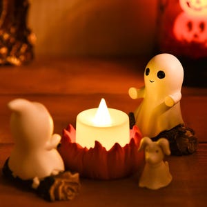 May include: A Halloween-themed display featuring a glowing LED candle in a red, petal-shaped holder, flanked by small, white ghost figurines and a dog figurine. The scene is set on a wooden surface, with a blurred background.