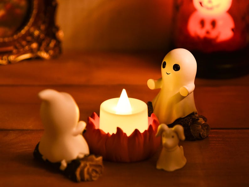 May include: A Halloween-themed display featuring a glowing LED candle in a red, petal-shaped holder, flanked by small, white ghost figurines and a dog figurine. The scene is set on a wooden surface, with a blurred background.