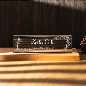 May include: A clear glass desk nameplate with a white floral design and the text "Kelly Cole Project Coordinator".