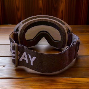 May include: Brown and black ski goggles with a wide strap. The strap has the word "BAY" printed in white. The goggles have a thick, padded frame and clear lenses. The goggles are designed for winter sports.