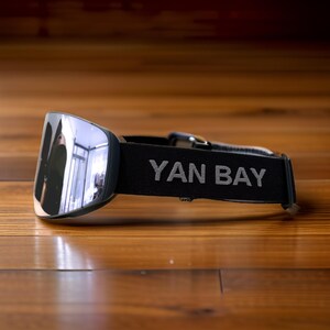 May include: Silver mirrored lens ski goggles with a black strap. The strap features the text "YAN BAY" in gray lettering. The goggles are resting on a wooden surface.
