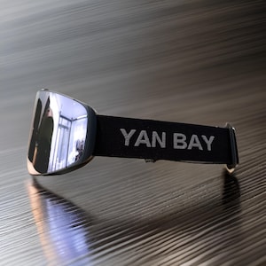 Può includere: Occhiali da sci neri con lente riflettente e cinturino nero con la scritta "YAN BAY" in bianco.